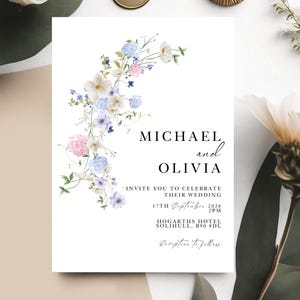 May include: A wedding invitation with a floral design. The invitation is white and features the names "MICHAEL and OLIVIA", along with the wedding details. The floral arrangement includes pastel blue, pink, and white flowers.