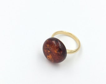 May include: A gold ring featuring a large, oval amber stone. The amber has a rich, warm brown color with intricate patterns. The ring's band is a simple, polished gold. A piece of jewelry.