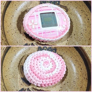 May include: A pink and white crocheted circle with a pink and white swirl pattern. The circle is sitting on a brown surface.