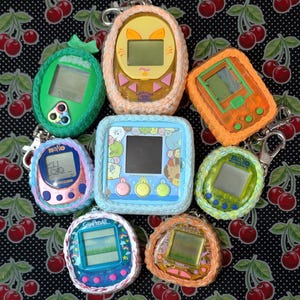 May include: Seven colorful crocheted Tamagotchi-style digital pet toys. The toys are in various colors, including orange, green, blue, pink, and yellow. Each toy has a small screen and buttons. The toys are all connected to keychains.