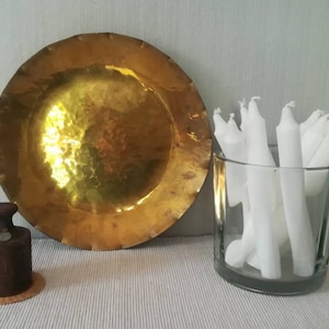 Könnte beinhalten: Eine große, gehämmerte Servierplatte aus Messing mit einem gewellten Rand, ein kleines Holzgewicht und eine Glasvase gefüllt mit weißen Kerzen.