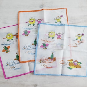 Könnte beinhalten: Drei weiße Taschentücher mit farbenfrohen Illustrationen und Bordüren. Jedes zeigt eine Cartoon-Sonne, Kinder, Wolken und fliegende Objekte. Die Bordüren sind rosa, orange und blau. Auf einer hellen Holzoberfläche.