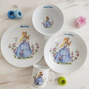 Könnte beinhalten: Ein weißes Porzellan-Kindergeschirr mit einem blauen und weißen Blumendesign, das ein Mädchen in einem blauen Kleid zeigt. Das Set enthält eine Schüssel, einen Teller und eine Tasse. Jedes Stück ist mit dem Namen "Marina" personalisiert.