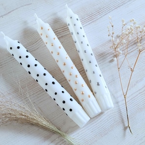 Könnte beinhalten: Drei weiße Spitzkerzen mit dekorativen Punktmustern. Eine Kerze hat schwarze Punkte, eine andere goldene Punkte und die dritte silberne Punkte. Die Kerzen sind auf einer hellen Holzoberfläche mit getrockneten floralen Akzenten angeordnet.