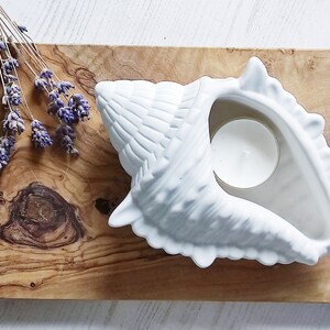 Könnte beinhalten: Weißer Kerzenhalter aus Keramik in Muschelform mit einer kleinen weißen Teelichtkerze im Inneren. Der Halter steht auf einer Holzoberfläche mit getrockneten Lavendelzweigen.