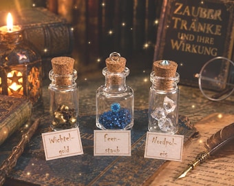 Könnte beinhalten: Drei kleine, durchsichtige Glasflaschen mit Korkstopfen. Die erste enthält goldfarbene Perlen mit der Aufschrift Wichtel-gold. Die zweite hat eine silberne Öse, blaue Perlen und ist mit feen-staub beschriftet. Die dritte enthält klare Kristalle mit der Aufschrift Nordpol-eis.