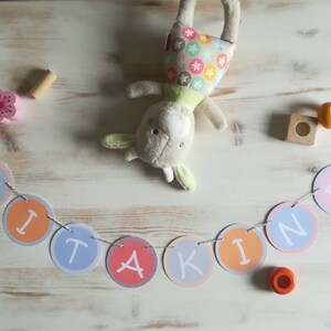 Könnte beinhalten: Ein farbenfrohes Banner mit dem Wort "KIND" in weißen Buchstaben auf einer Reihe von pastellfarbenen Kreisen. Das Banner ist mit weißem Garn befestigt und hat rosa Blumenakzente.