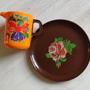Könnte beinhalten: Ein brauner Plastikteller mit einer einzelnen rosa Rose und grünen Blättern, die darauf gedruckt sind. Neben dem Teller steht ein kleiner orangefarbener Plastikkrug mit einem Blumendesign.