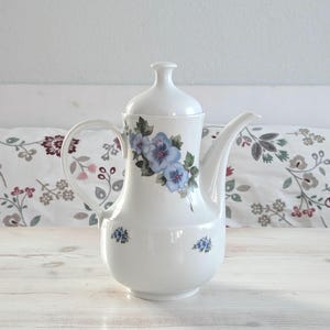 Könnte beinhalten: Weiße Kaffeekanne aus Keramik mit Deckel, Henkel und Ausguss. Die Kanne ist mit blauen Blumenmustern verziert. Der Hintergrund umfasst ein floral gemustertes Kissen und einen weißen Holztisch.
