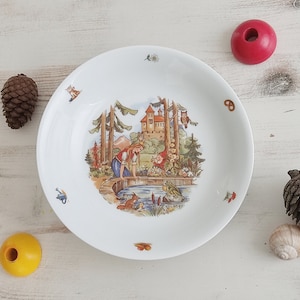 Könnte beinhalten: Ein weißer Keramikteller mit einer bunten Illustration einer Märchenszene mit einem Schloss, einem Mädchen, einem Zwerg und einem Frosch. Der Teller ist mit kleinen Details wie Blumen, Bäumen und Vögeln verziert.