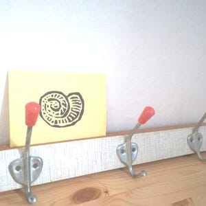 Könnte beinhalten: Ein Holz-Kleiderständer mit drei Haken. Die Haken sind silberfarben mit roten Plastikspitzen. Eine gelbe Notizkarte mit einer schwarz-weißen Illustration einer Muschel lehnt an dem Ständer.