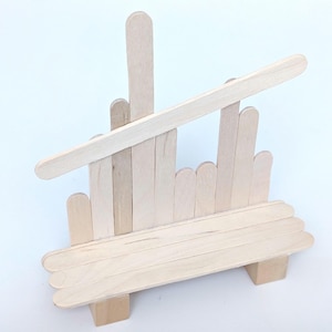 Könnte beinhalten: Eine Miniatur-Staffelei aus Holz, hergestellt aus Bastelstäbchen. Die Staffelei hat eine Basis, eine Rückenstütze und ein schräges Oberteil. Die natürliche Holzfarbe sorgt für eine schlichte, rustikale Ästhetik. Ideal für die Präsentation kleiner Kunstwerke oder Fotos.