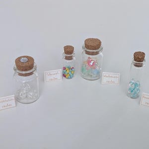 Könnte beinhalten: Vier kleine Glasflaschen mit Korken, jede mit bunten Perlen und Kristallen gefüllt. Kleine weiße Karten mit deutschem Text sind vor jeder Flasche platziert. Die Flaschen sind auf einer weißen Oberfläche angeordnet.
