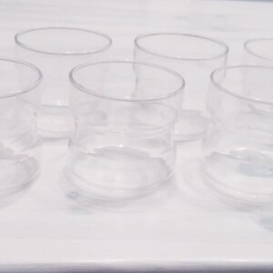 Könnte beinhalten: Sechs klare Glasbecher mit leicht ausgestelltem Rand. Die Becher sind in einem Halbkreis auf einer weißen Oberfläche angeordnet.