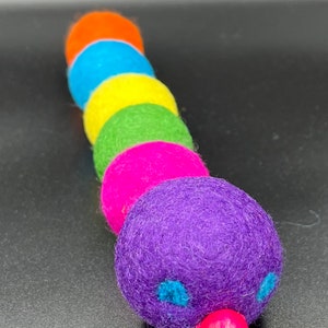 Puede incluir: Un juguete de oruga de fieltro colorido con una cabeza morada, ojos azules y una nariz rosa. El cuerpo est&aacute; hecho de seis bolas de fieltro en los colores del arco&iacute;ris.