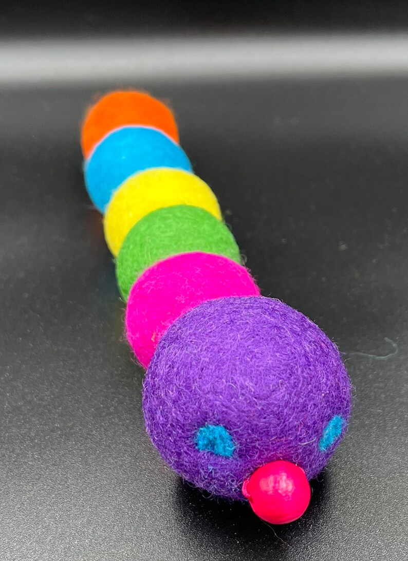 Puede incluir: Un juguete de oruga de fieltro colorido con una cabeza morada, ojos azules y una nariz rosa. El cuerpo est&aacute; hecho de seis bolas de fieltro en los colores del arco&iacute;ris.