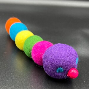 Puede incluir: Un juguete de oruga de fieltro colorido con una cabeza morada, una nariz rosa y ojos azules. La oruga est&aacute; hecha de bolas de fieltro de color naranja, azul, amarillo, verde y rosa.