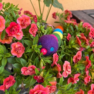 Puede incluir: Un juguete de oruga de fieltro colorido con una cabeza morada y ojos rojos, anidado en una maceta de flores rosas.