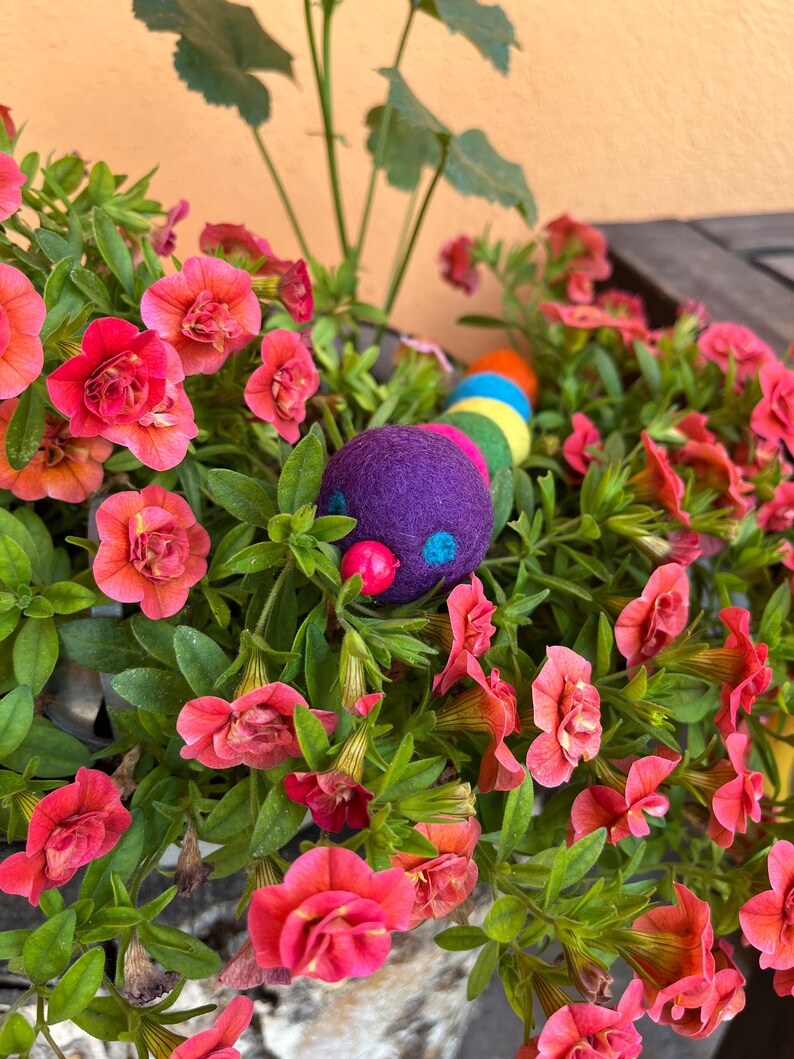 Puede incluir: Un juguete de oruga de fieltro colorido con una cabeza morada y ojos rojos, anidado en una maceta de flores rosas.