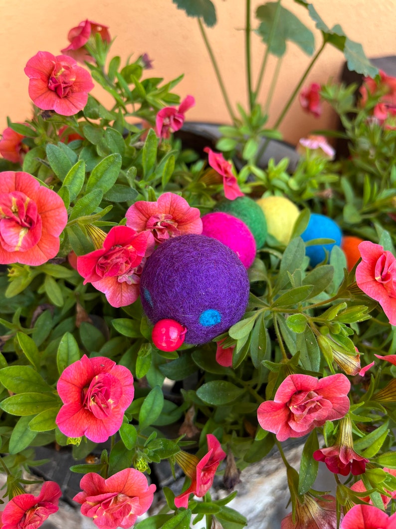 Puede incluir: Un juguete de oruga de fieltro colorido con una cabeza morada y una nariz roja, rodeado de flores rosas.