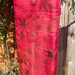Pode incluir: Um lenço retangular de seda num tom vermelho vibrante, com um padrão disperso de impressões botânicas de folhas em castanho escuro. O tecido tem uma textura suave e ligeiramente refletora com formas de folhas naturais visíveis.