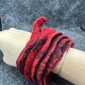 Puede incluir: Una bufanda larga y estrecha hecha de lana afieltrada con un patrón de color moteado en rojo y negro vibrante. La tela tiene una apariencia texturizada y difusa con bordes irregulares, envuelta varias veces en espiral para crear un aspecto en capas.