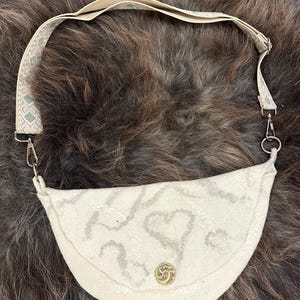May include: Cream-colored felt crossbody bag with a decorative heart pattern in gray. The bag features a gold-colored button closure and a patterned beige strap with silver-tone hardware. The bag is designed to be worn over the shoulder.