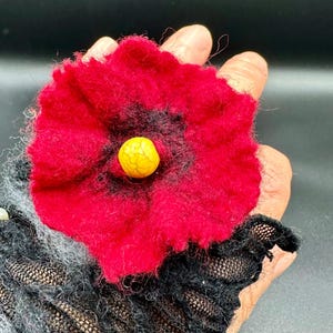 Könnte beinhalten: Eine rote Filzblume mit gelbem Zentrum, getragen an einem schwarzen und grauen Armband.