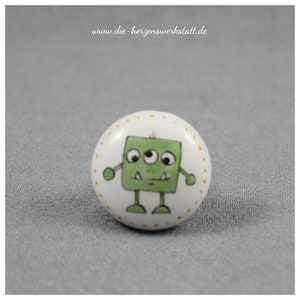 Puede incluir: Un pomo de cerámica blanca con una ilustración de un monstruo verde de tres ojos. El monstruo tiene una cabeza cuadrada, brazos y piernas. El pomo está decorado con pequeños puntos naranjas y el texto "www.die-herzenswerkstatt.de".