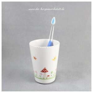 Puede incluir: Portacepillos de dientes de cer&aacute;mica blanca con un dise&ntilde;o caprichoso que presenta setas rojas y blancas, mariposas y flores. El portacepillos tiene un cepillo de dientes azul en su interior.