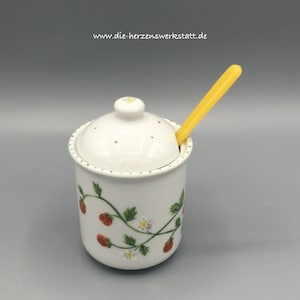 Könnte beinhalten: Weiße Keramik-Zuckerdose mit Deckel und gelbem Plastiklöffel. Die Dose hat ein Erdbeer-Design mit grünen Blättern und weißen Blüten.