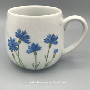 Op de afbeelding: Witte keramische mok met een afgeronde vorm en een groot handvat. De mok is versierd met blauwe korenbloem illustraties en groene stelen. Het websiteadres "www.die-herzenswerkstatt.de" staat onderaan afgedrukt.