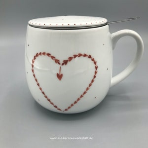 Peut inclure: Une tasse à thé en céramique blanche avec un motif de cœur rouge et un couvercle en acier inoxydable. La tasse a une poignée et une petite passoire à l'intérieur. Le texte "www.die-herzenswerkstatt.de" est imprimé au bas de l'image.