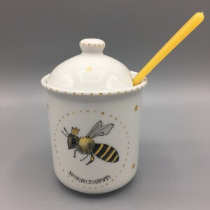Puede incluir: Tarro de miel de cerámica blanca con un diseño de abeja dorada y la palabra "summsumm" en la parte delantera. El tarro tiene una tapa y viene con un cucharón de miel amarillo.