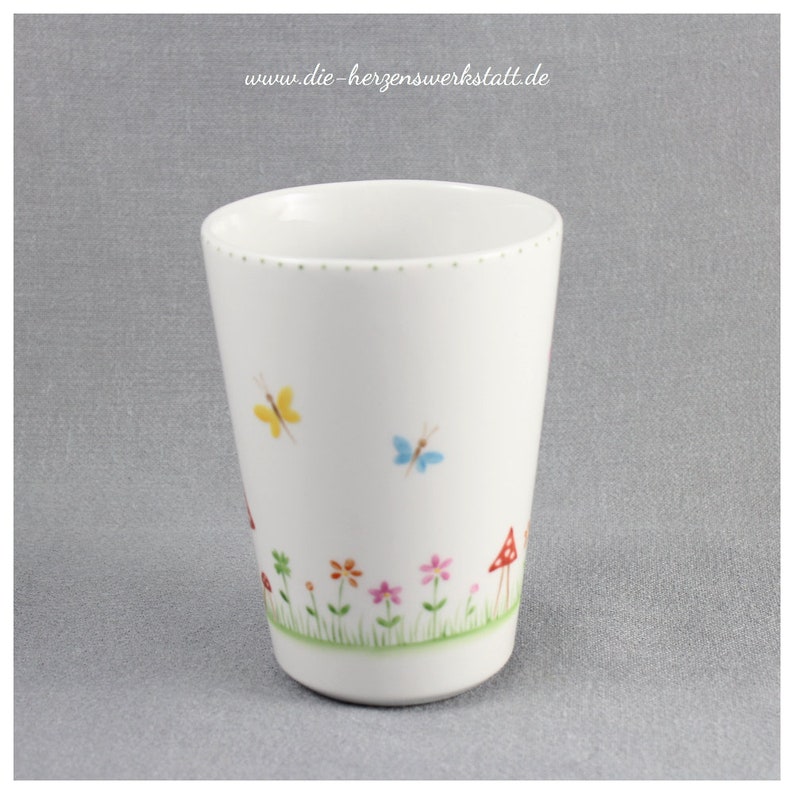 Puede incluir: Taza de cer&aacute;mica blanca con un dise&ntilde;o caprichoso que presenta mariposas, flores y setas. La taza tiene un borde dorado y una l&iacute;nea punteada alrededor de la parte superior.