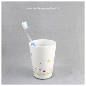 Puede incluir: Portacepillos de dientes de cer&aacute;mica blanca con un dise&ntilde;o floral con mariposas, flores y un hongo. Un cepillo de dientes azul est&aacute; en el portacepillos.