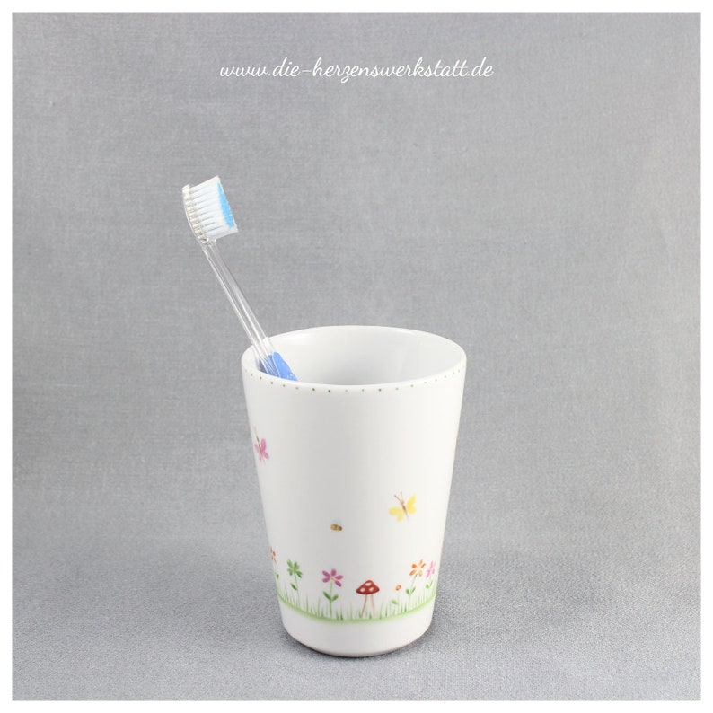 Puede incluir: Portacepillos de dientes de cer&aacute;mica blanca con un dise&ntilde;o floral con mariposas, flores y un hongo. Un cepillo de dientes azul est&aacute; en el portacepillos.