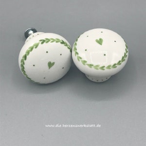 Op de afbeelding: Twee witte keramische knoppen met groene bladeren en witte stippen. Elke knop heeft een klein hartje erop geschilderd.