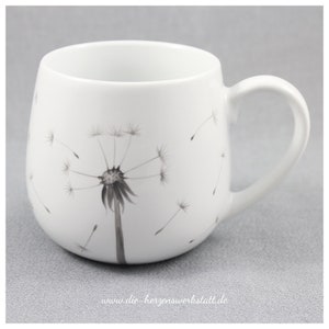 Pode incluir: Caneca de cerâmica branca com um desenho de dente-de-leão preto e branco. A caneca tem um corpo arredondado e uma alça. O texto "www.die-herzenswerkstatt.de" está impresso na parte inferior da caneca.