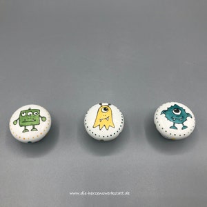 Peut inclure: Trois boutons en céramique avec des motifs de monstres. Les boutons sont blancs avec des accents verts, jaunes et bleus. Les monstres ont respectivement trois yeux, un seul œil et deux yeux.