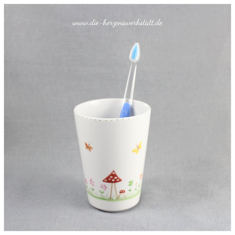 Puede incluir: Portacepillos de dientes de cer&aacute;mica blanca con un dise&ntilde;o caprichoso que presenta setas rojas y blancas, mariposas y flores. El portacepillos tiene un cepillo de dientes azul en su interior.