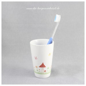 Puede incluir: Portacepillos de cer&aacute;mica blanca con un dise&ntilde;o de hongo y mariposa. El portacepillos tiene un cepillo de dientes azul en su interior.