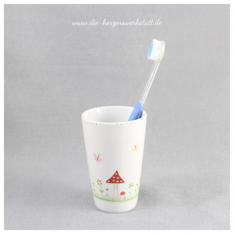 Puede incluir: Portacepillos de cer&aacute;mica blanca con un dise&ntilde;o de hongo y mariposa. El portacepillos tiene un cepillo de dientes azul en su interior.