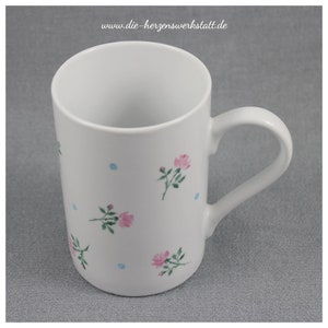 Könnte beinhalten: Weißer Keramikbecher mit gebogenem Henkel, verziert mit rosa Rosenillustrationen und kleinen blauen Punkten. Auf dem Becher steht der Text "www.die-herzenswerkstatt.de".