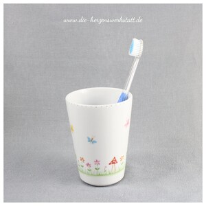 Puede incluir: Portacepillos de dientes de cer&aacute;mica blanca con un dise&ntilde;o floral con mariposas, flores y setas. El portacepillos tiene un cepillo de dientes azul en su interior.