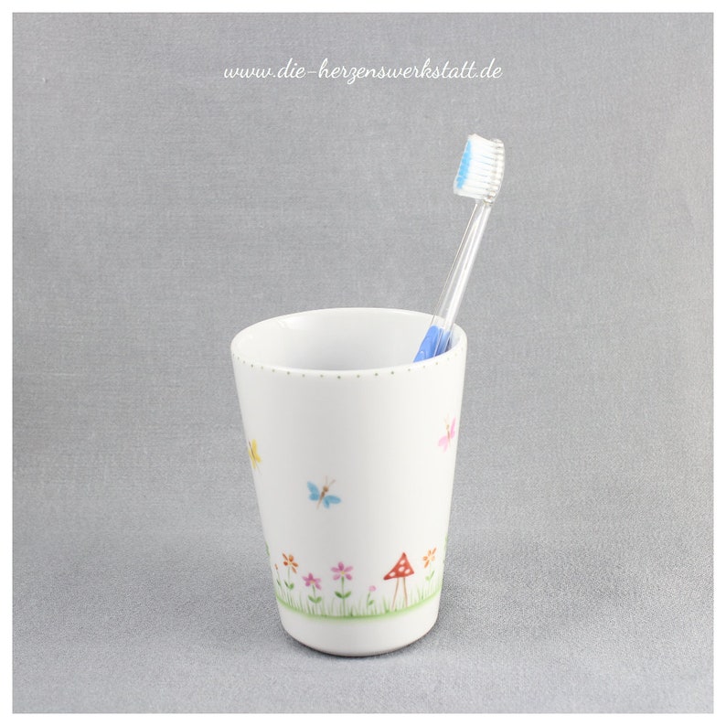 Puede incluir: Portacepillos de dientes de cer&aacute;mica blanca con un dise&ntilde;o floral con mariposas, flores y setas. El portacepillos tiene un cepillo de dientes azul en su interior.