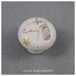 Puede incluir: Pomo de cerámica blanco con un diseño de unicornio pintado a mano. El unicornio tiene un cuerpo rosa, una crin arcoíris y un cuerno dorado. El pomo está decorado con estrellas doradas y puntos de colores.