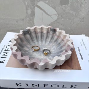 Könnte beinhalten: Eine dekorative Schale mit gewelltem Rand in Rosa, Grau und Weiß, die zwei Ringe enthält. Ein Ring ist goldfarben mit Perlen, der andere silberfarben mit einem goldenen Herz. Die Schale liegt auf einem Stapel Bücher.