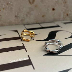 Könnte beinhalten: Zwei abstrakt geformte Ringe, einer in Gold und einer in Silber. Die Ringe sind aus Metall und haben ein einzigartiges, organisches Design.