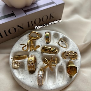 Könnte beinhalten: Eine weiße Keramik-Ring-Schale mit goldenen Ringen, darunter ein Ring mit einem Seestern-Design, ein Ring mit einem Ketten-Design und ein Ring mit der Gravur "You Are My Sunshine".